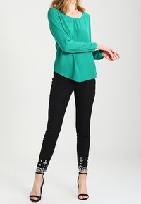 Blusa de seda verde azulado con mangas largas, combinada con pantalones ajustados negros que presentan bordados de flores blancas en los tobillos y tacones negros de punta abierta.