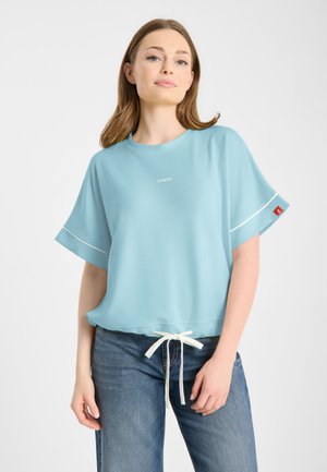 Vrouw draagt een lichtblauw loszittend kort mouw shirt met witte trekkoord en spijkerbroek, staand tegen een effen witte achtergrond.