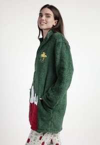 Joven mujer sonriendo, vestida con una chaqueta verde de felpa con un parche de pájaro amarillo y un diseño de dibujos animados en rojo y blanco, combinada con leggings blancos con un patrón.