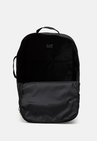 Musta reppu, jossa on vetoketjullinen avaus, sileä nailonmateriaali ja logo sisävuorausmateriaalissa. Sisus on tilava ja tummasta kankaasta.