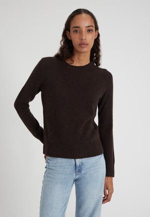 Vrouw met krullend haar in een donkerbruine lange mouwen trui en lichtblauwe jeans, staand tegen een effen lichte achtergrond.
