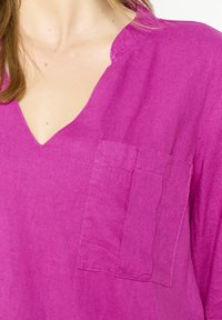 Fuchsiová ľanová košeľa s výstrihom do V, dlhými rukávmi a náprsné vrecko. Tkanina má hladkú textúru s jemným leskom.