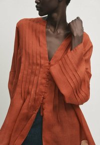 Rustfarvet button-up skjorte lavet af letvægtsstof. Har lodrette plisseringer, en dyb V-hals og brede ærmer med en afslappet pasform.