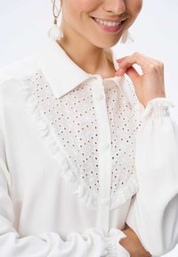 Biała bluzka z kołnierzykiem, zapięciem na guziki oraz delikatnym wstawką z koronką i haftem. Bufiaste rękawy z marszczonym mankietem.