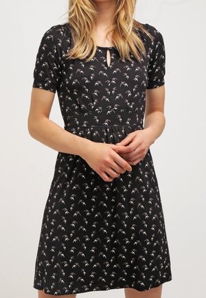 Femme portant une robe noire à manches courtes avec un petit motif d'oiseaux et un décolleté en trou de serrure, les mains légèrement jointes devant.