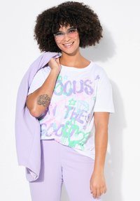Hvid T-shirt med farverigt, abstrakt grafisk design bestående af pink, grønne og lilla former. Kombinationen er sat sammen med lyse lilla bukser og jakke.