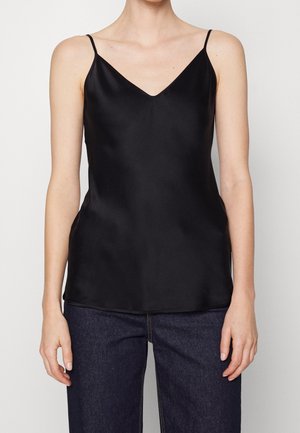 Persona con una camiseta sin mangas de satén negro con tirantes finos y vaqueros azul oscuro, de pie frente a un fondo claro y liso.