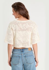 Un haut court en tricot de couleur crème, à coupe ample, présentant un délicat motif floral ajouré et des manches larges. Porté avec un short en jean taille haute.
