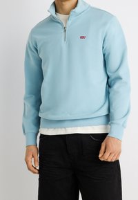 Homme portant un sweat-shirt zippé bleu clair avec un petit logo rouge Levi's sur la poitrine, un pantalon noir et une chemise blanche en dessous.