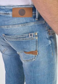 Jeans de mezclilla azul claro con corte recto, que presentan un parche de cuero marrón, acentos en los bolsillos y detalles en costura rojos y blancos.