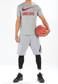 Camiseta gris de Nike con "CAVALIERS" en rojo, pantalones cortos grises, mallas de compresión negras y zapatillas negras, sosteniendo un baloncesto.