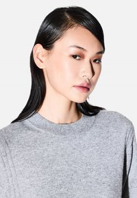 Jonge vrouw met steil zwart haar, een neuspiercing en neutrale make-up, draagt een grijze trui tegen een eenvoudige witte achtergrond.