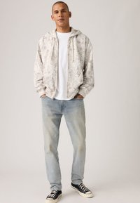 Φούτερ με κουκούλα σε ανοιχτό γκρι χρώμα με υφασμάτινο, διακοσμητικό μοτίβο· συνδυασμένο με ανοιχτόχρωμο μπλε τζιν και μαύρα αθλητικά παπούτσια με λευκές σόλες.