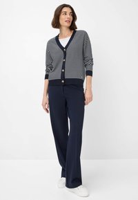 Marineblau-gestreifter Cardigan mit V-Ausschnitt und goldenen Knöpfen, kombiniert mit dunkelblauen weit geschnittenen Hosen und weißen Sneakers. Weißer Hintergrund.