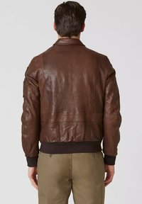 Uomo che indossa una giacca di pelle marrone e pantaloni khaki, visto da dietro su uno sfondo semplice.