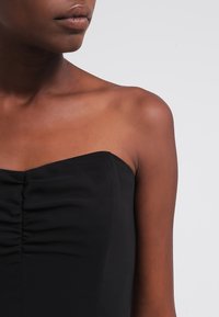 Černý bezramínkový top s nařasenou texturou na hrudi. Hladká tkanina s přiléhavým střihem, který ukazuje čistý výstřih a ramena.