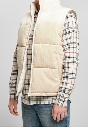 Hombre con chaleco sin mangas beige acolchado sobre camisa de cuadros blanca, roja y negra con botones y vaqueros azules sobre fondo neutro.