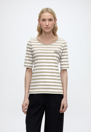 Blonde vrouw met los haar, gekleed in een beige-wit gestreept T-shirt met korte mouwen en de tekst "Tutto bene!", en een zwarte broek, staat neutraal.