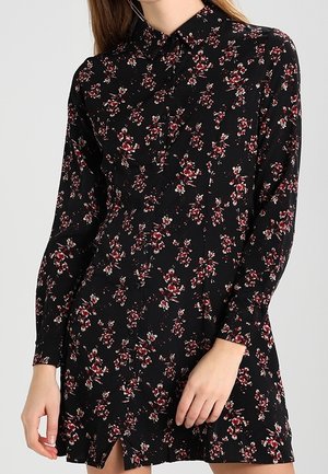 Sort langærmet kjole med rødt og hvidt blomsterprint, knaplukning foran og en krave, båret af en person med én hånd hvilende nær kanten.