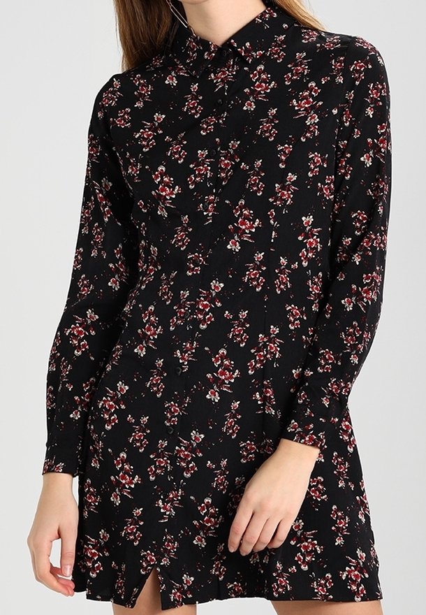 Robe noire à manches longues avec motif floral rouge et blanc, boutonnée sur le devant, avec un col, portée par une personne avec une main posée près de l'ourlet.
