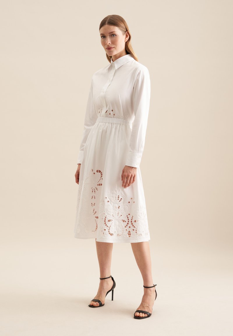 Weißes Hemdkleid mit ausgeschnittener Blumenstickerei am Saum, Knopfverschluss vorne, Kragen, elastischem Bund und hohen Sandalen mit Knöchelriemen.