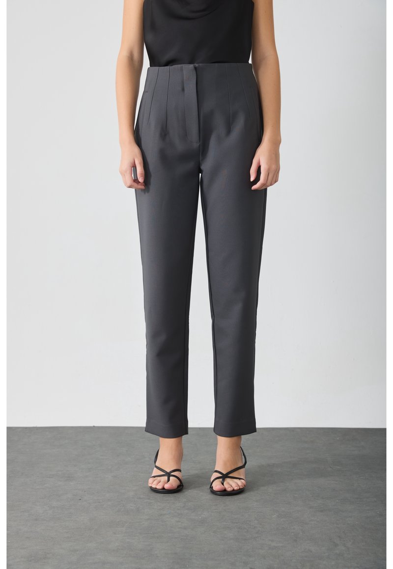 Personne portant un pantalon gris foncé sur mesure et un haut noir sans manches, debout sur un sol gris contre un mur blanc, sandales noires à brides visibles.