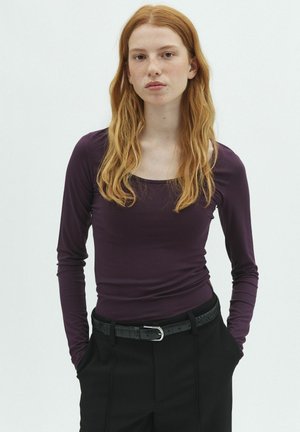 Jeune femme aux longs cheveux roux portant un t-shirt ajusté à manches longues violet foncé et un pantalon noir avec une ceinture noire, les mains dans les poches.