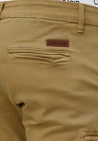 Kaki hlače izrađene od mekog materijala s stražnjim džepom i smešnim kožnim detaljem s logotipom. Ravna prednja strana, glatka tekstura.