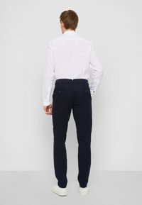 Marineblaue Hose mit geradem Schnitt, aus Baumwollmaterial gefertigt, ausgestattet mit zwei hinteren Taschen und einem Reißverschluss, kombiniert mit einem weißen Hemd.