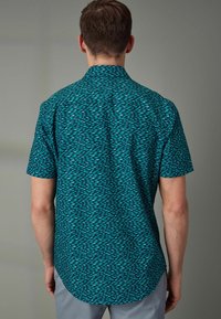 Camicetta a maniche corte realizzata in tessuto leggero, con uno sfondo turchese e un motivo geometrico blu scuro, e un orlo curvo.