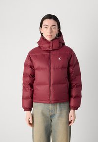 Giubbotto imbottito bordeaux con colletto alto, chiusura con zip e logo del marchio. Presenta un design trapuntato e polsini elastici. Indossato sopra un jeans.