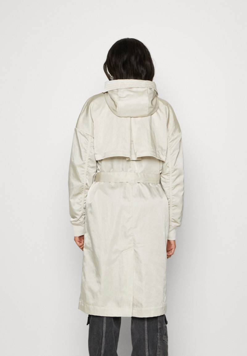 Gabardina larga de color beige claro con capucha, sección superior recortada, puños elásticos y cintura con cinturón, con una textura suave y brillante.