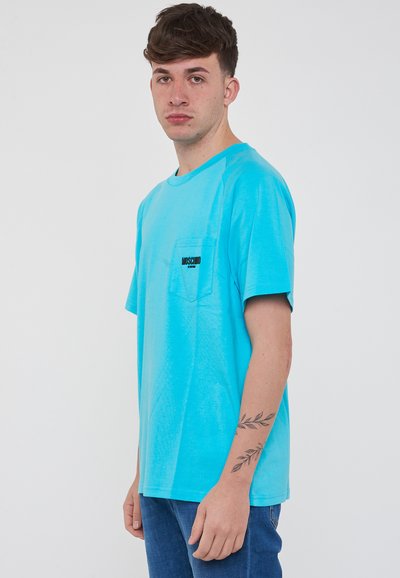 T-shirt di cotone turchese con collo rotondo, maniche corte e un piccolo logo a tasca nero. Design semplice, vestibilità rilassata.