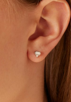 Zeskantige studs in goudkleurig metaal, met een witte steen en een kleine cluster van drie heldere rhinestones aan de basis.