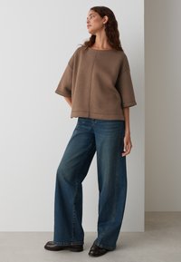 Sweatshirt oversize marron avec un décolleté large et des manches courtes, associé à un jean en denim bleu ample et des bottines marron. Texture lisse.
