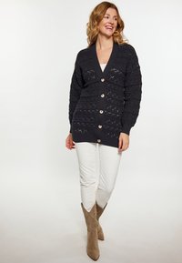 Cardigan lavorato a maglia di un blu scuro con scollo a V profondo, caratterizzato da motivi testurizzati e grandi bottoni di legno, abbinato a pantaloni bianchi e stivali color cammello.