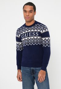 Il maglione di colore blu navy presenta un colletto decorato in bianco e beige, con la scritta "GUESS" in evidenza. Maniche lunghe con polsini e orlo a costine.