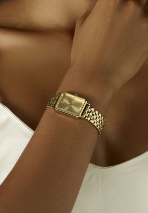 Montre rectangulaire en or avec un cadran texturé et un bracelet tissé doré. Portée sur un poignet nu, avec des index horaires minimalistes et sans chiffres.