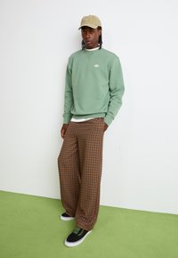 Mintgrön sweatshirt med rund hals, bruna rutiga vida byxor och svarta sneakers med gröna snören; mot en vit bakgrund.