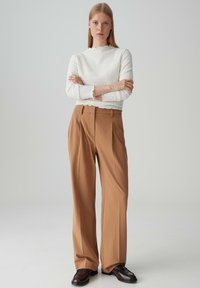 Witte getextureerde blouse met gegolfde randen, gecombineerd met bruine hooggeknipte geplooide broek, afgewerkt met donkere schoenen. Neutrale achtergrond.