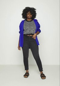 Camisa oversized azul, top cropped com padrão de ondas preto e branco, jeans pretos e sandálias pretas. Apresenta cabelo encaracolado e óculos de sol redondos.