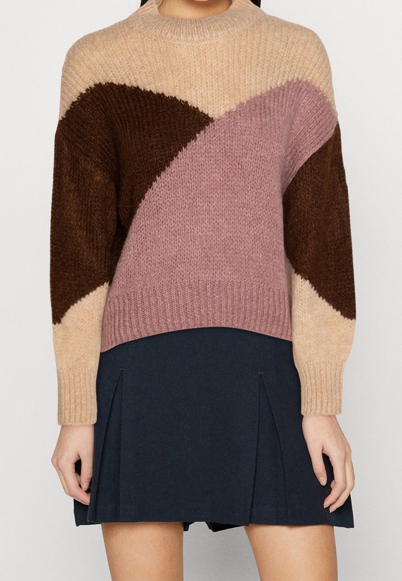 Femme portant un pull à blocs de couleur beige, marron et mauve, assorti d'une jupe plissée bleu marine, les mains détendues le long du corps.