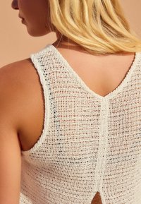 Hvid strikket tanktop med et løst, tekstureret design. Har en v-halsudskæring og en åben ryg med en lille slids. Let og åndbart materiale.