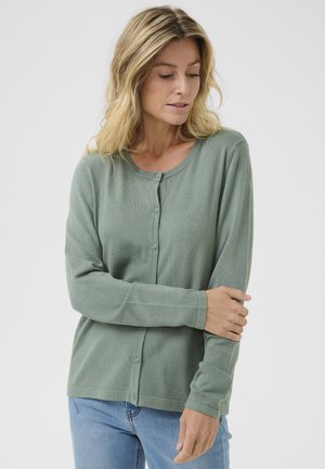 Cardigan vert en tissu doux, avec un décolleté rond, des manches longues, une fermeture à boutons et une texture légèrement côtelée.