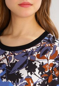 Blusa de estampado floral con un escote de canalé negro, que presenta colores azul, naranja y blanco sobre un tejido ligero.