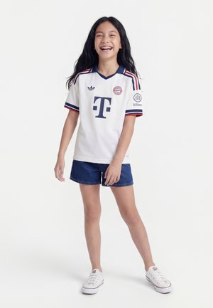 Ung pige smilende, iført hvid Bayern München fodboldtrøje, blå shorts og hvide sneakers, står foran en ensfarvet baggrund.