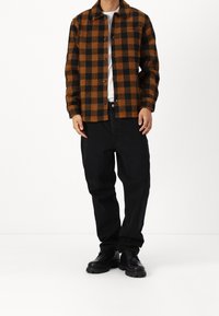 Hnědá a černá kostkovaná flanelová košile s knoflíkovou légou vpředu, nošená přes bílou košili, doplněná černými kalhotami a černými šněrovacími botami.