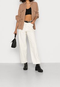 Bruine overhemdd met twee borstzakken, zwarte korte tanktop, witte wijde broek, zwarte enkelboots en een kleine zwarte handtas.