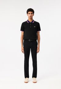 Zwarte polo met rode en blauwe accenten, korte mouwen, groen logo, gecombineerd met zwarte broek en beige sneakers. Eenvoudig, aangesloten ontwerp.
