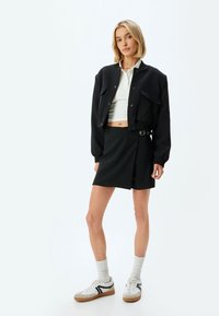 Zwart cropped jack met twee voorzakken, witte polo, zwarte wikkelrok en witte sneakers met grijze accenten en zwarte strepen.